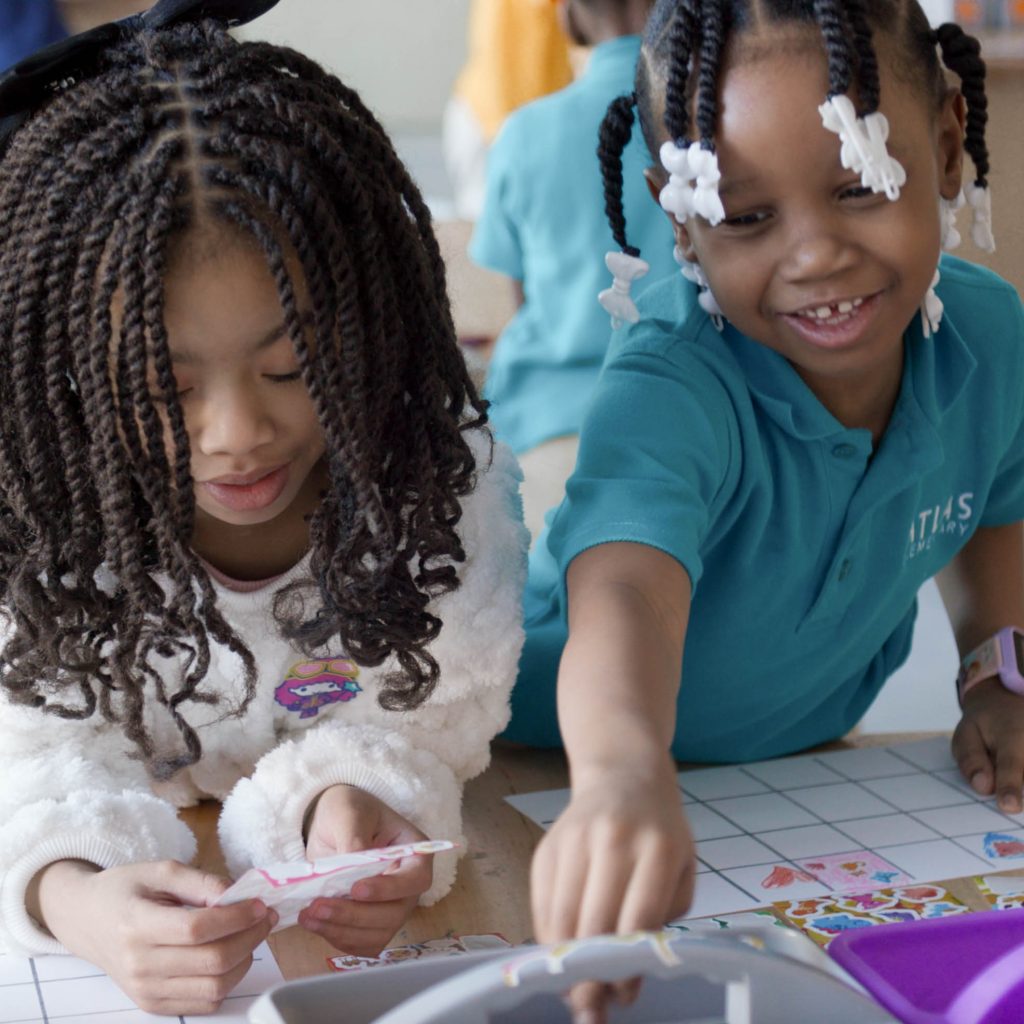 Two young girls work on a paper craft at a tiled table, cutting and sticking colored shapes and stickers.